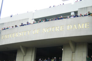 'Notre Dame' Stadium Facade with People Looking Over Edges, with 'University of Notre Dame' Engraved in Gold Lettering